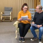 Gallery 1 - A white woman and a white man sit together on a small bench reading from a script they're both holding in their hands